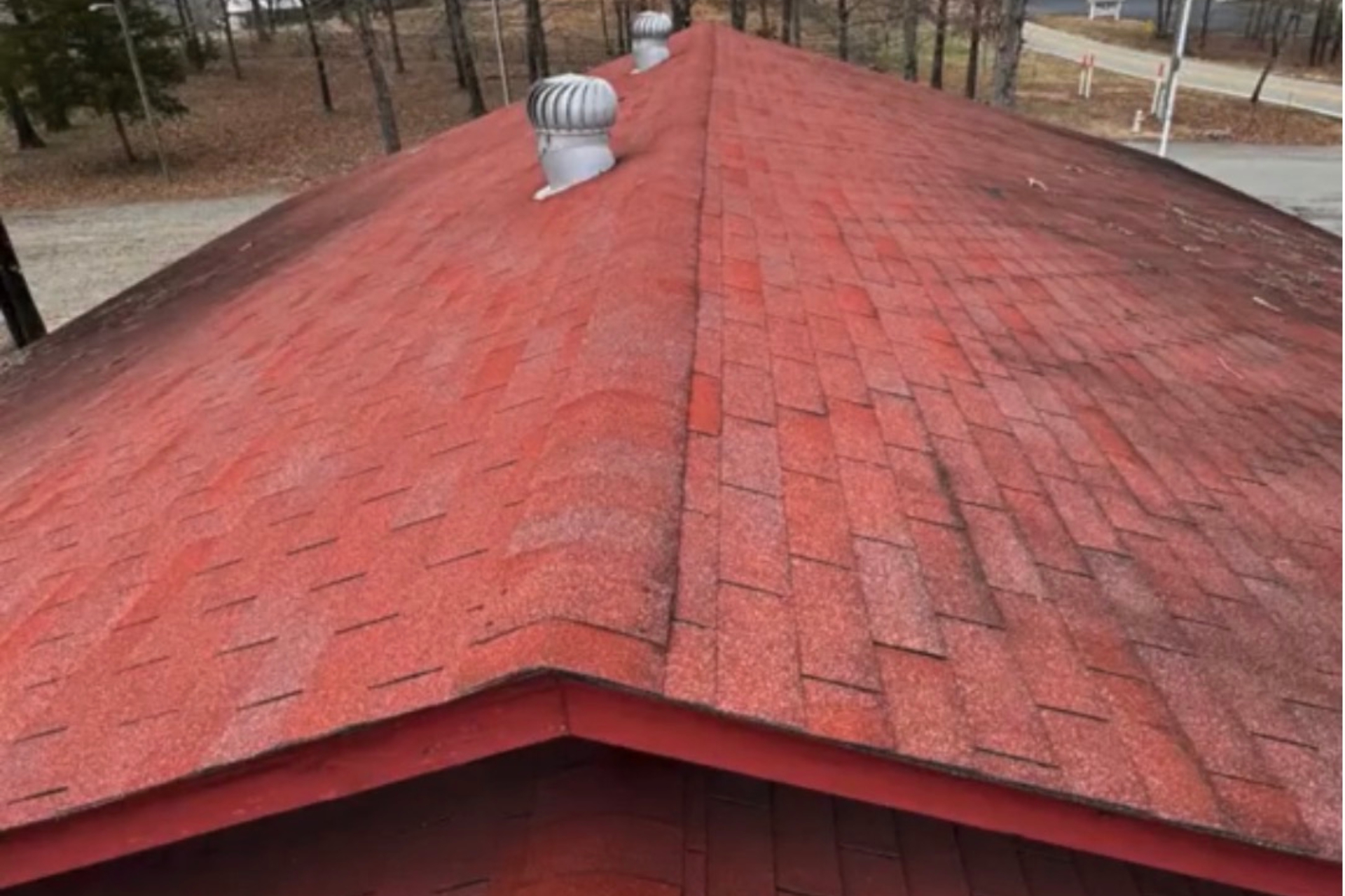 Red shingled roof with two silver turbine roof vents, surrounded by leafless trees and a road visible in the background.