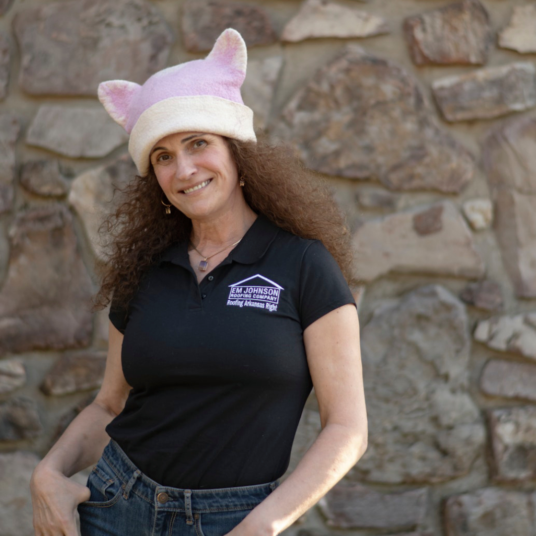 A woman with curly brown hair wears a pink and white cat ear hat, a black polo shirt with an EJ Johnson name tag, and blue jeans, standing in front of a stone wall and smiling at the camera.