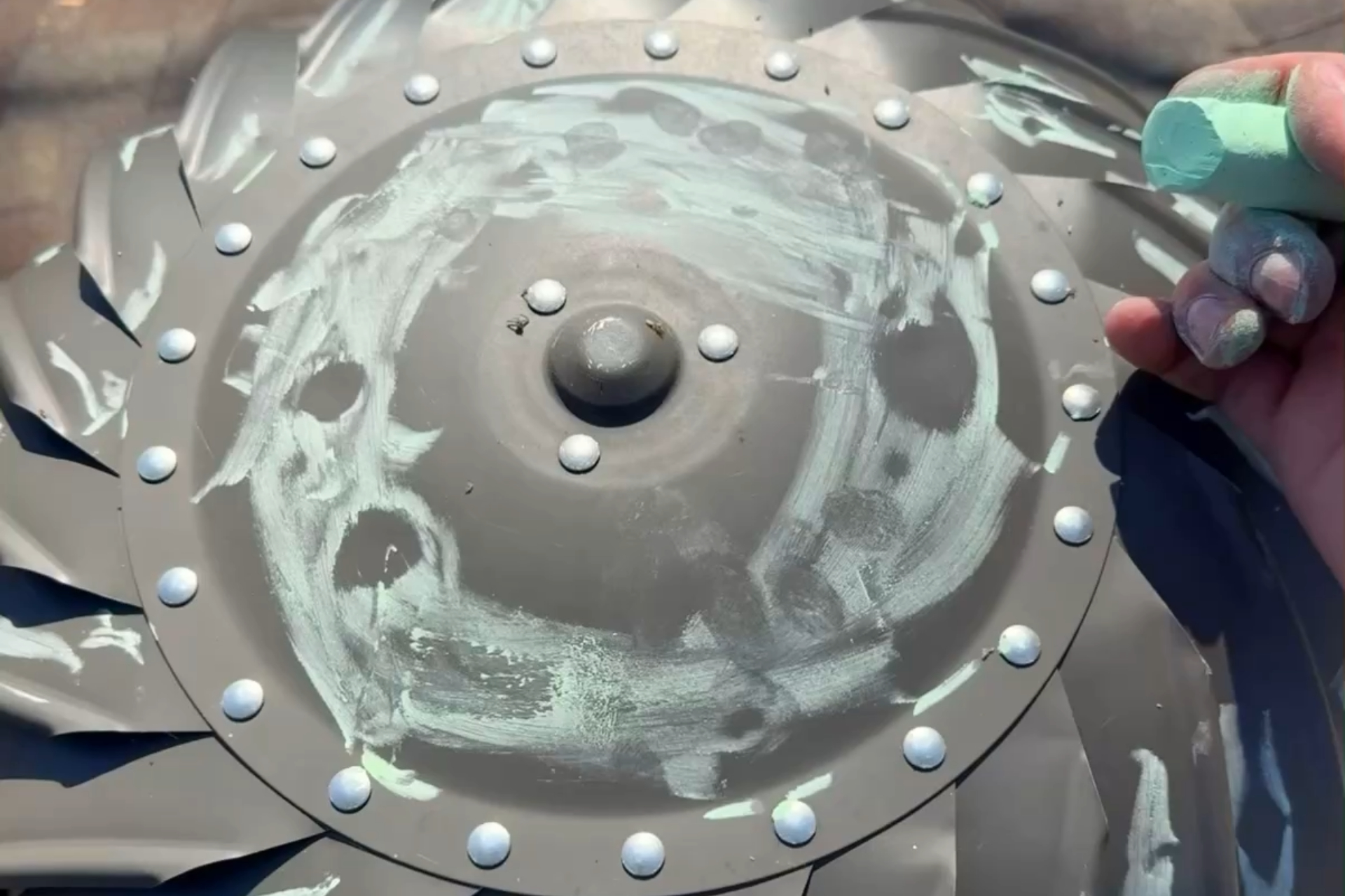 A hand holding green chalk beside a round, metal surface with bolts, partially covered in green chalk marks and fingerprints.