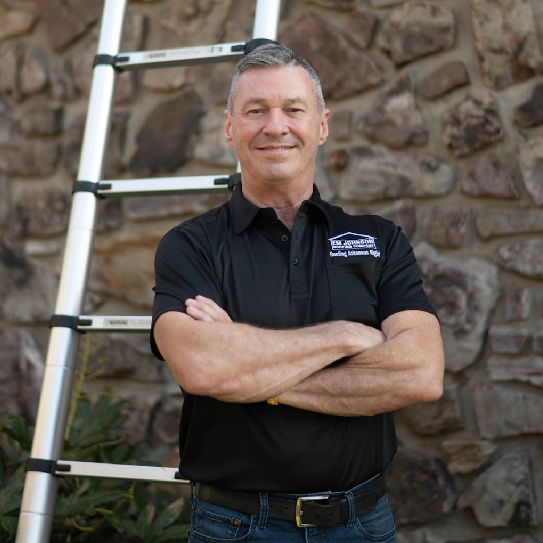 A man in a black polo shirt stands in front of a stone wall and a ladder, smiling with his arms crossed. Some greenery is visible near his side.