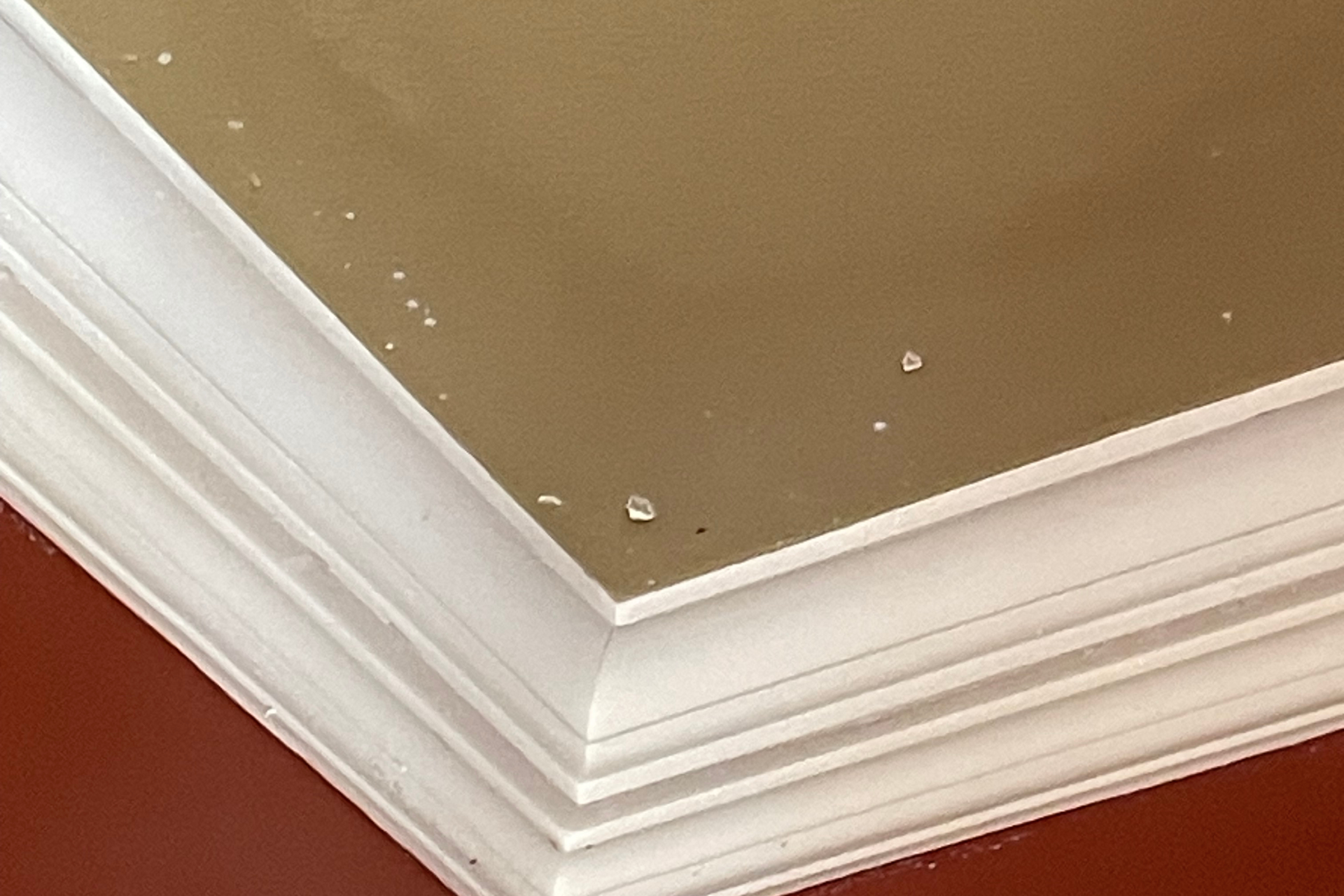 Close-up of a ceiling corner with beige and red walls, white crown molding, and some chipped paint and small holes visible on the beige ceiling surface.