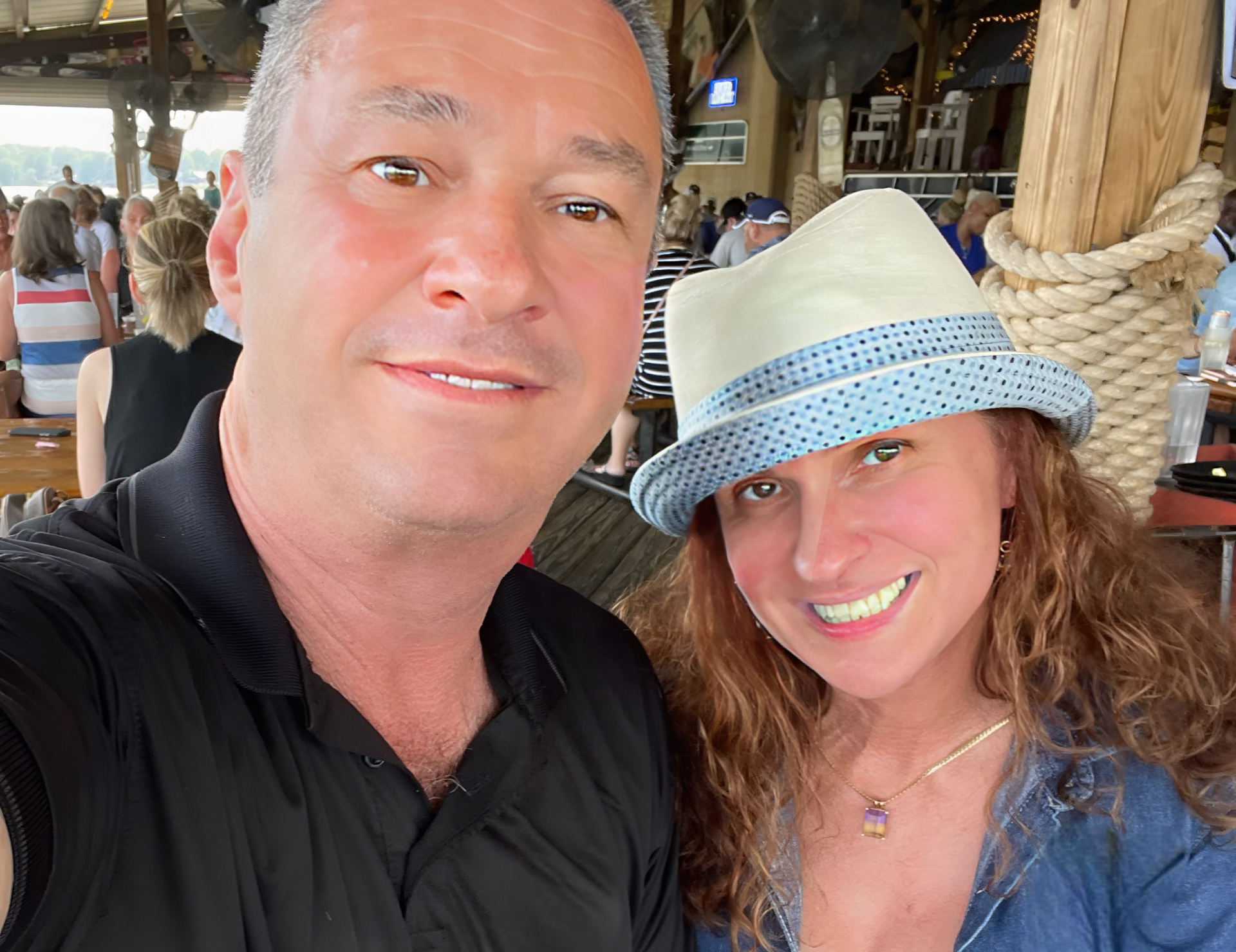 A man in a black shirt and a woman in a blue shirt and light-colored hat smile together for a selfie at a busy outdoor venue with wooden posts and people in the background.