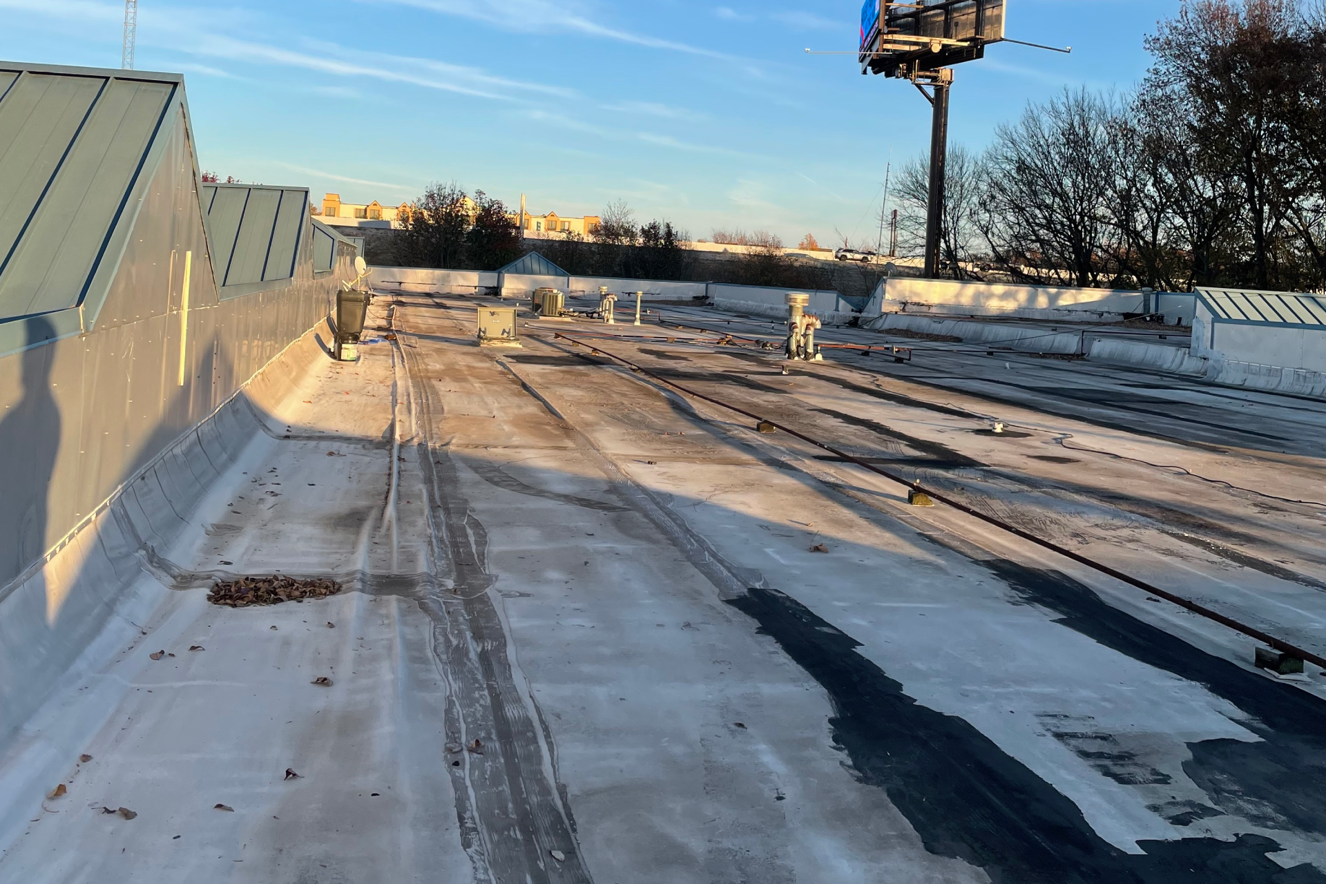 A flat commercial roof with a white membrane coating, scattered debris, rooftop equipment, and some shadows; trees and a billboard are visible in the background under a clear blue sky.